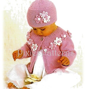 Könnte beinhalten: Eine rosa gestrickte Babyjacke mit weißen Blumenverzierungen und eine passende rosa gestrickte Mütze mit weißen Blumenverzierungen.