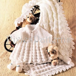 May include: A white knitted baby outfit with a cardigan, a bonnet, booties, and a matching blanket. The outfit is laid out on a wooden surface with two teddy bears.