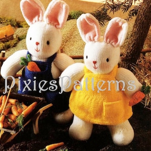 May include: Two knitted white bunny rabbits wearing clothes. One bunny is wearing blue overalls and holding a carrot. The other bunny is wearing a yellow dress and holding a carrot. The bunnies are sitting on a brown surface with a wheelbarrow full of carrots in the background.