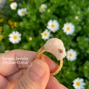 May include: A gold ring featuring a carved pale pink lion head with a small red gemstone eye. The band has a textured, etched surface. The text Serene Jewelry Collections is visible on the left side of the image in a white cursive font.