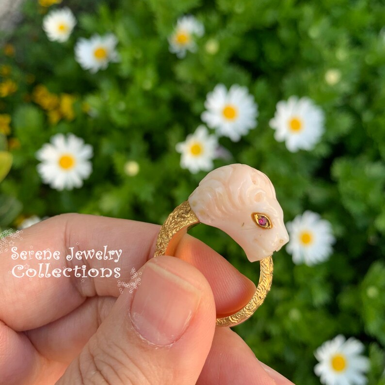 May include: A gold ring featuring a carved pale pink lion head with a small red gemstone eye. The band has a textured, etched surface. The text Serene Jewelry Collections is visible on the left side of the image in a white cursive font.