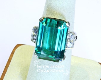 May include: A ring featuring a large, rectangular, emerald-cut teal gemstone set in a silver-toned metal band. The central stone is flanked on each side by two smaller, round, clear stones. The text Serene Jewelry Collections is visible below.