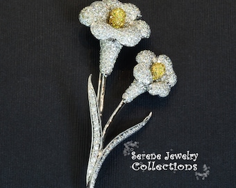 May include: A silver brooch featuring two white flowers with yellow centers, set with sparkling diamonds. The flowers are arranged on a stem with leaves, creating a delicate and elegant design.