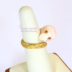 May include: A gold ring featuring a textured band and a carved pale pink stone lion head ornament with two small red gemstone eyes. The text Serene Jewelry Collections is visible in purple script on the display stand behind the ring.