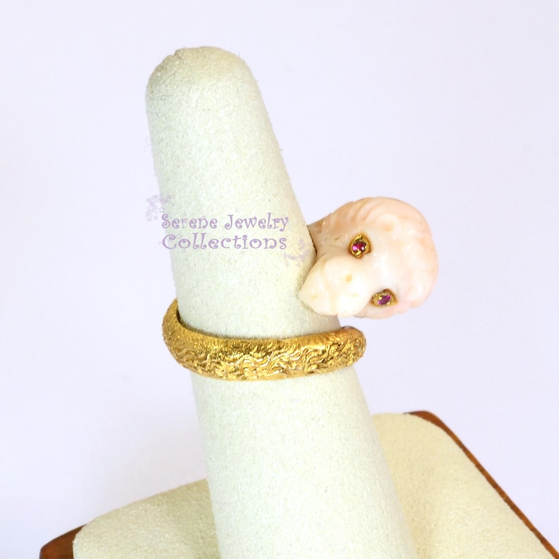 May include: A gold ring featuring a textured band and a carved pale pink stone lion head ornament with two small red gemstone eyes. The text Serene Jewelry Collections is visible in purple script on the display stand behind the ring.