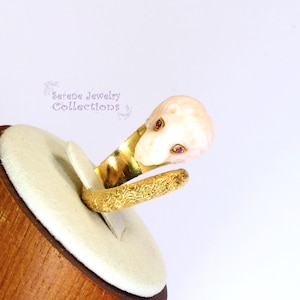 May include: A gold ring featuring a textured band and a carved pale pink lion head ornament with two small red gemstone eyes. The text Serene Jewelry Collections is visible in the background. The ring is displayed on a rounded beige stand.