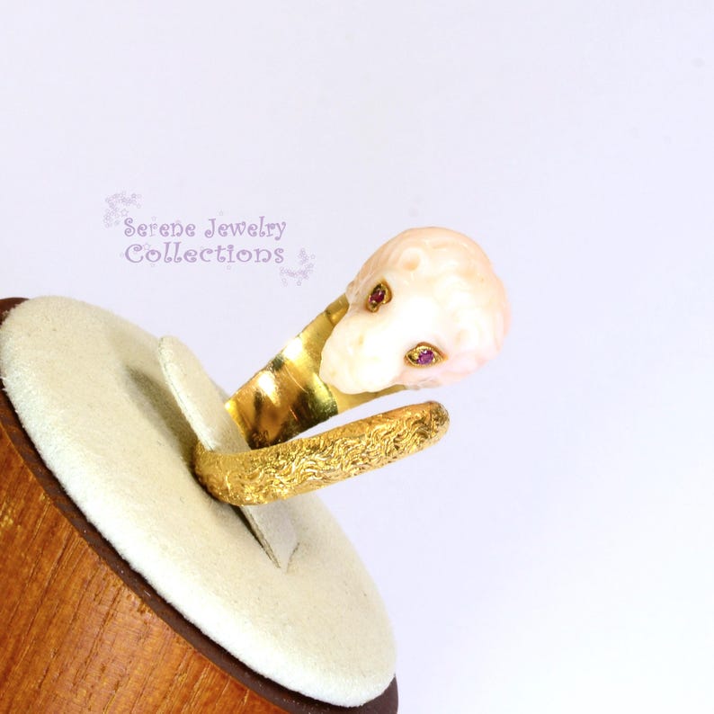 May include: A gold ring featuring a textured band and a carved pale pink lion head ornament with two small red gemstone eyes. The text Serene Jewelry Collections is visible in the background. The ring is displayed on a rounded beige stand.