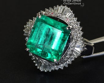 May include: A large emerald green gemstone ring with a square cut stone surrounded by a halo of smaller diamonds. The ring is set in white gold and has a delicate, intricate design.