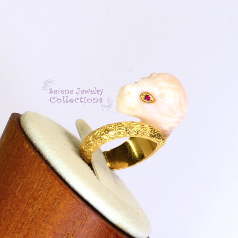 May include: A gold ring featuring a textured band and a carved pale pink stone lion head ornament. The lion has a small, vibrant red gemstone set as an eye. The text Serene Jewelry Collections is visible in the background in a light purple font.