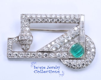 May include: A silver and diamond brooch with a geometric design and a green emerald stone in the center. The brooch is set in a rectangular frame with a diamond-encrusted border.