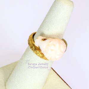 May include: A gold ring featuring a textured, ornate band and a carved, pale pink floral-shaped stone centerpiece. The stone is accented with a small, embedded red gemstone. The text Serene Jewelry Collections is visible near the ring base.