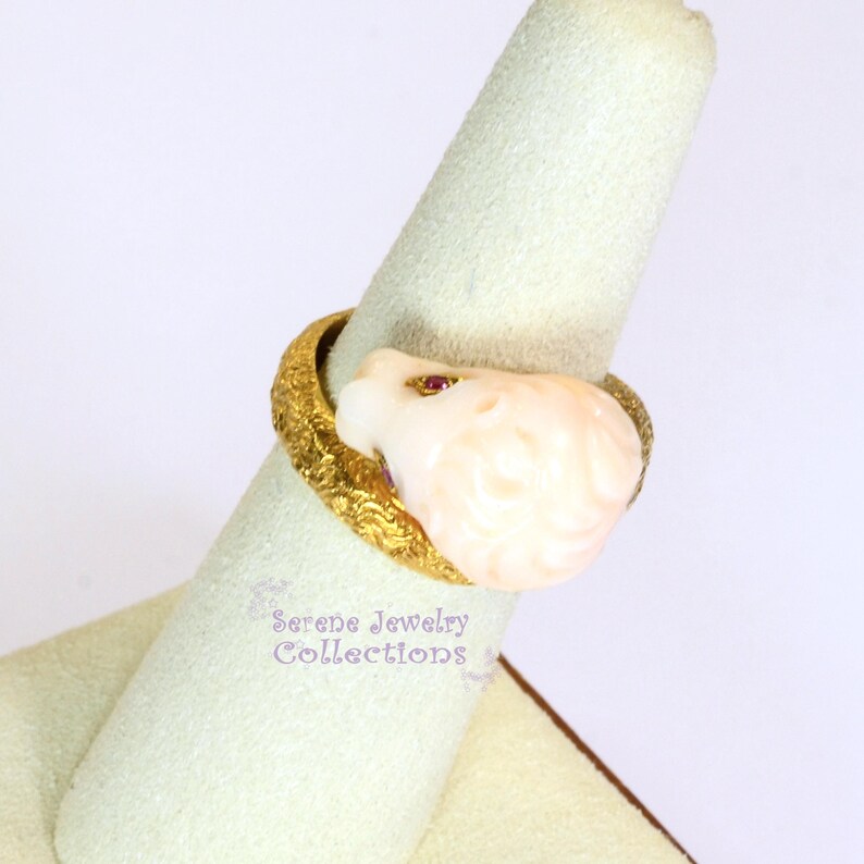 May include: A gold ring featuring a textured, ornate band and a carved, pale pink floral-shaped stone centerpiece. The stone is accented with a small, embedded red gemstone. The text Serene Jewelry Collections is visible near the ring base.