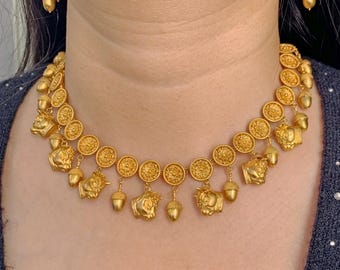 Könnte beinhalten: Eine goldfarbene Halskette mit einer Kette aus runden Medaillons mit Blumenmustern, durchsetzt mit baumelnden Anhängern in Form von Eicheln und stilisierten Tierköpfen. Passende Ohrhänger mit Eichelmotiven vervollständigen das Set.