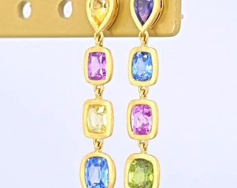 May include: A pair of gold drop earrings featuring four vertically aligned, bezel-set gemstones each. The left earring displays yellow, pink, yellow, and blue stones. The right earring features purple, blue, pink, and green stones in a gold setting.