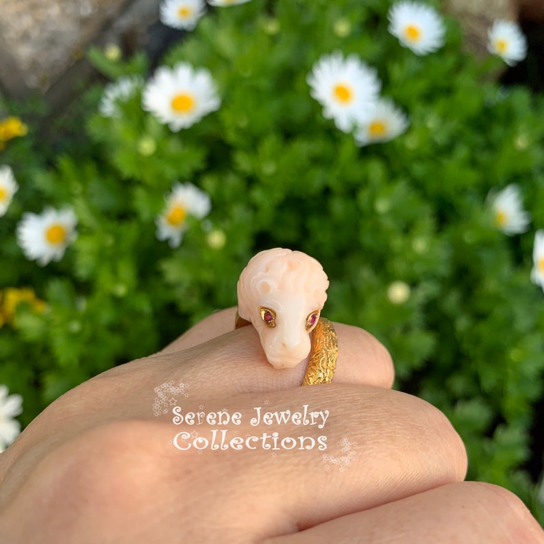 May include: A gold ring featuring a carved pale pink lion head with small red gemstone eyes. The band has a textured, organic finish. The text "Serene Jewelry Collections" is printed in white on the lower portion of the image.