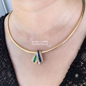 May include: A gold necklace with a pendant featuring three rows of baguette-cut gemstones. The top row is green, the middle row is blue, and the bottom row is clear. The necklace is on a model's neck.