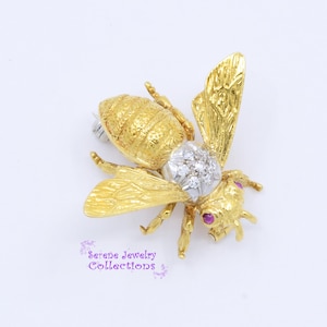 May include: A gold bee brooch featuring a textured abdomen, wings with detailed vein patterns, a thorax set with clear round stones, and two small pink circular stones for eyes. The text Serene Jewelry Collections is printed in purple.