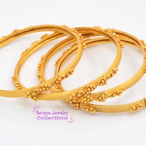 May include: A set of four gold-toned bangles featuring a ribbed texture and small, clustered bead accents spaced evenly around the circumference. The text Serene Jewelry Collections is printed in purple at the bottom of the image.