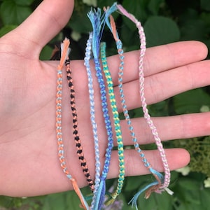 May include: A collection of eight friendship bracelets in various colors, including pink, blue, green, yellow, and orange. The bracelets are made of braided string and have frayed ends.