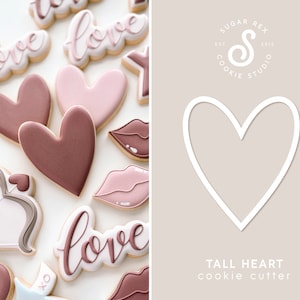 May include: A cookie cutter in the shape of a tall heart. The cutter is white and is shown on a light brown background. The text "TALL HEART cookie cutter" is below the cutter.