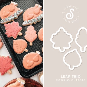 May include: A set of three cookie cutters shaped like leaves. The cutters are made of white metal and are arranged on a light gray background. The text "LEAF TRIO cookie cutters" is printed below the cutters.