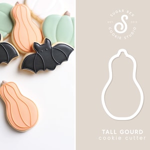 May include: A cookie cutter in the shape of a tall gourd, with the text "TALL GOURD cookie cutter" below it. The cutter is white and is shown against a light brown background. The logo "SUGAR REX COOKIE STUDIO EST 2013" is shown in the top right corner.