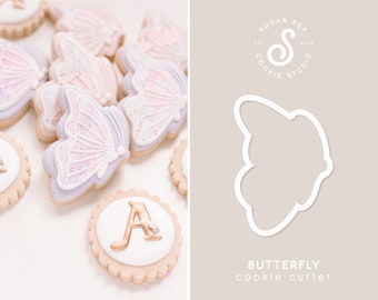 May include: A set of butterfly-shaped sugar cookies decorated with purple and pink icing. The cookies are arranged on a white surface. A cookie cutter in the shape of a butterfly is shown on a light brown background. The text "BUTTERFLY cookie cutter" is below the cookie cutter.