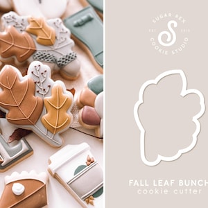 May include: A cookie cutter in the shape of a bunch of fall leaves. The cutter is white and is on a light brown background. The text "FALL LEAF BUNCH cookie cutter" is below the cutter.