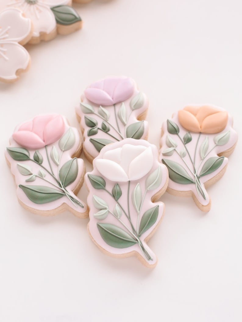 May include: Five decorated sugar cookies with floral designs. Each cookie features a different coloured flower with green leaves. The flowers are pink, purple, white, and orange.