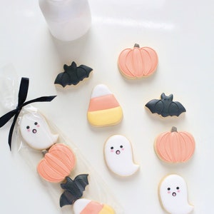 May include: Halloween-themed cookies in various shapes, including ghosts, pumpkins, bats, and candy corn. The cookies are decorated with pastel colors and packaged in a clear cellophane bag tied with a black ribbon. A white bottle with a black and white striped straw is also present.