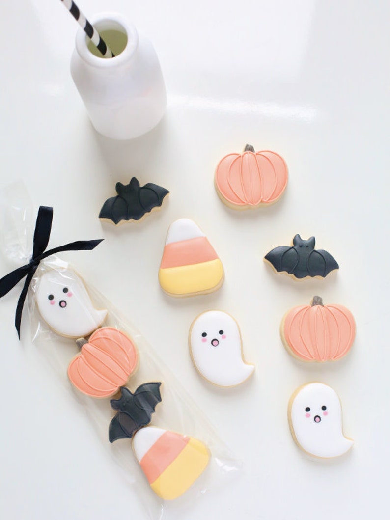 May include: Halloween-themed cookies in various shapes, including ghosts, pumpkins, bats, and candy corn. The cookies are decorated with pastel colors and packaged in a clear cellophane bag tied with a black ribbon. A white bottle with a black and white striped straw is also present.