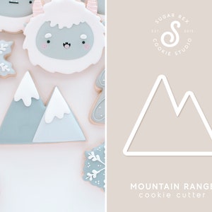 May include: A white cookie cutter shaped like a mountain range. The cookie cutter is on a light brown background with the text "MOUNTAIN RANGE cookie cutter" below it.