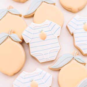 K&ouml;nnte beinhalten: Eine Sammlung von dekorierten Zuckerkeksen mit Zitronenmotiven und einem Baby-Strampler mit einem Zitronenmotiv. Die Kekse sind mit blauem und wei&szlig;em Zuckerguss verziert.