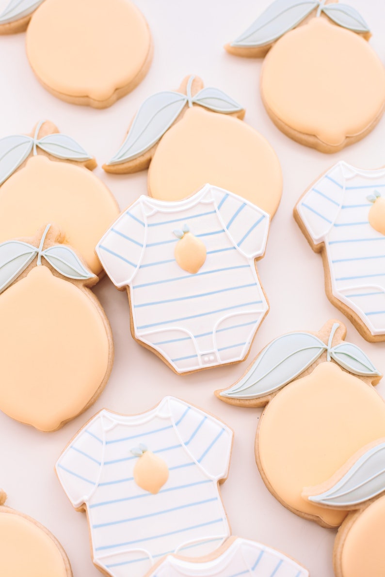K&ouml;nnte beinhalten: Eine Sammlung von dekorierten Zuckerkeksen mit Zitronenmotiven und einem Baby-Strampler mit einem Zitronenmotiv. Die Kekse sind mit blauem und wei&szlig;em Zuckerguss verziert.