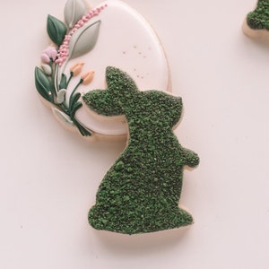 May include: A white and green sugar cookie decorated with a green bunny silhouette and a floral design with pink and yellow accents.