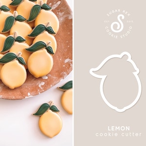 May include: A close-up of a cookie cutter in the shape of a lemon. The cookie cutter is white and is on a light gray background. The text "LEMON cookie cutter" is below the cookie cutter.