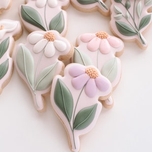 K&ouml;nnte beinhalten: Dekorierte Zuckerkekse mit Blumenmotiven. Die Kekse zeigen wei&szlig;e, rosa und lila Bl&uuml;ten mit gr&uuml;nen Bl&auml;ttern.