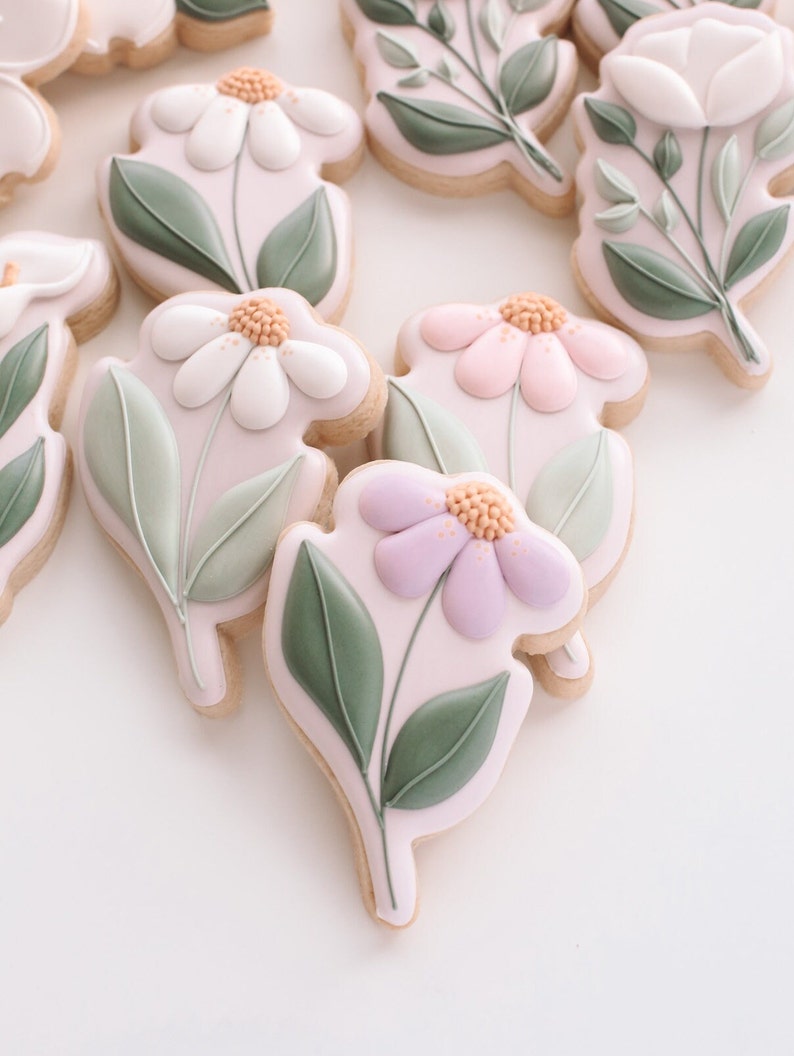 K&ouml;nnte beinhalten: Dekorierte Zuckerkekse mit Blumenmotiven. Die Kekse zeigen wei&szlig;e, rosa und lila Bl&uuml;ten mit gr&uuml;nen Bl&auml;ttern.