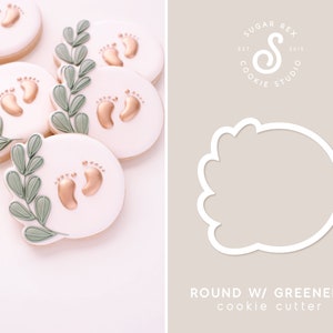 May include: A set of five oval-shaped sugar cookies decorated with gold baby footprints and green leaves. The cookies are arranged in a pile on a white surface. The image also shows a white cookie cutter in the shape of a round cookie with a scalloped edge. The text "ROUND W/ GREENERY cookie cutter" is printed below the cookie cutter.