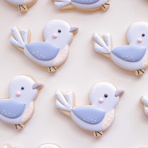 May include: A collection of nine blue and white bird-shaped sugar cookies decorated with pink and white details. The cookies are arranged in a pattern on a white background.