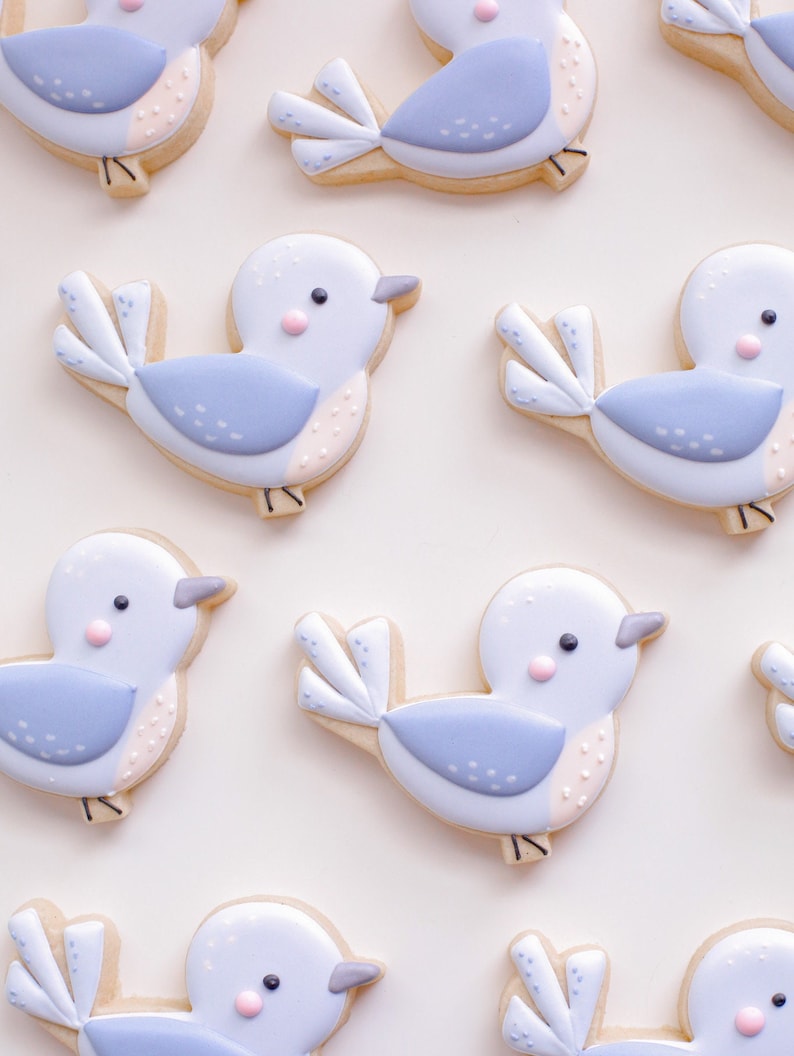 May include: A collection of nine blue and white bird-shaped sugar cookies decorated with pink and white details. The cookies are arranged in a pattern on a white background.