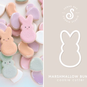 May include: A cookie cutter in the shape of a marshmallow bunny. The cutter is white and is shown on a light brown background. The text "MARSHMALLOW BUNNY cookie cutter" is below the cutter.