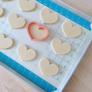 May include: A baking sheet with a blue silicone baking mat. The mat has a grid pattern and numbers printed on it. There are 10 heart-shaped cookies on the mat, and a pink heart-shaped cookie cutter.