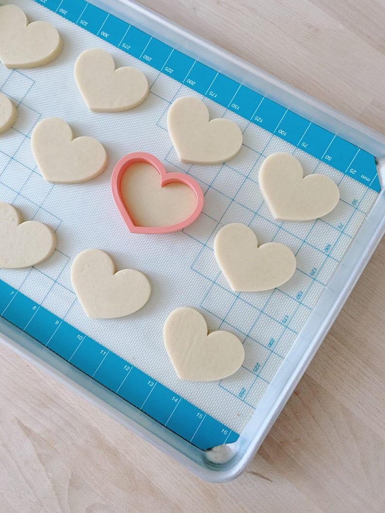 May include: A baking sheet with a blue silicone baking mat. The mat has a grid pattern and numbers printed on it. There are 10 heart-shaped cookies on the mat, and a pink heart-shaped cookie cutter.
