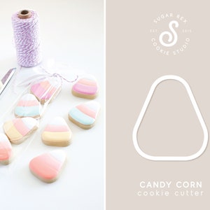 May include: Candy corn-shaped cookies in a clear cellophane bag tied with a white ribbon. The cookies have layers of pink, yellow, and blue icing. A white candy corn cookie cutter is next to the cookies. A spool of pink and white twine and gold scissors are also in the image.