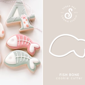 May include: A pink and green fish bone cookie cutter with a white outline. The cutter is shaped like a fish bone with a rounded head and tail. The text "FISH BONE cookie cutter" is printed below the cutter. The text "SUGAR REX COOKIE STUDIO EST 2015" is printed in the top right corner of the image.