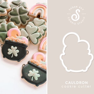May include: A black and green cookie cutter shaped like a cauldron with a rounded top and a handle. The cookie cutter is on a light gray background with the text "CAULDRON cookie cutter" below it.