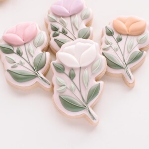 May include: Four decorated sugar cookies with floral designs. Each cookie features a single flower with green leaves and stems. The flowers are pink, white, light purple, and orange.