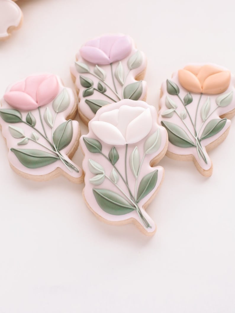 May include: Four decorated sugar cookies with floral designs. Each cookie features a single flower with green leaves and stems. The flowers are pink, white, light purple, and orange.