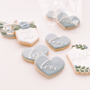 May include: Assortment of decorated sugar cookies with blue and white icing. The cookies feature the words "love", "bride to be", and "miss to mrs".